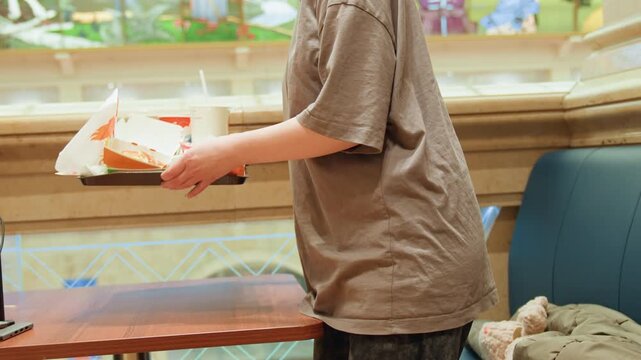 Young person carries tray across table, casual customer in tshirt picking up takeout meal from booth at mall food court, boxed burger and drink visible, ambient chatter and warm interior, candid