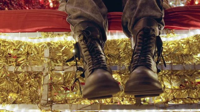 Boots dangling above golden tinsel, closeup of soldier sitting on storefront ledge with vintage leather combat boots, warm holiday lights, slow pensive movement, cinematic grain and cozy evening mood