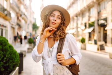 A smiling tourist with a backpack and hat enjoys a sunset stroll. The woman poses on a city street...
