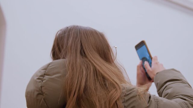 Young woman rear view with phone beneath festive ceiling, puffer coat and long hair framed by glowing lights and ornate architecture content creator captures upward scene, scrolling and smiling