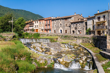Traditional stone houses lining a small river with cascades in Sant Feliu de Pallerols, Catalonia, scenic mountain village surrounded by green hills © StockMediaSeller
