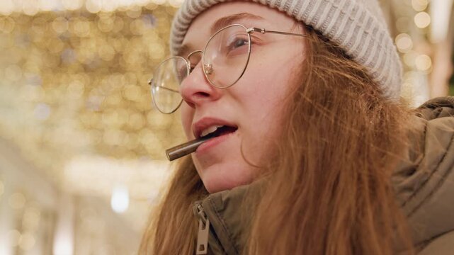White woman chewing pen under lights, round glasses and knit hat framing focused expression while journaling lyrics in winter market, closeup captures thoughtful mood, creative process and soft