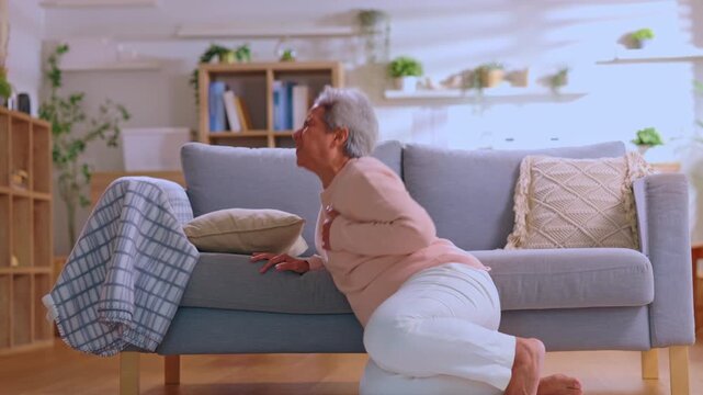 Senior woman suffers sudden heart attack feeling severe chest pain falling onto floor near comfortable sofa inside bright living room experiencing deep distress needing urgent medical help