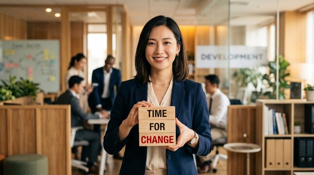 Businesswoman stands in a modern office holding a wooden block sign reading Time for Change while coworkers talk in the background among desks, plants, and glass partitions.