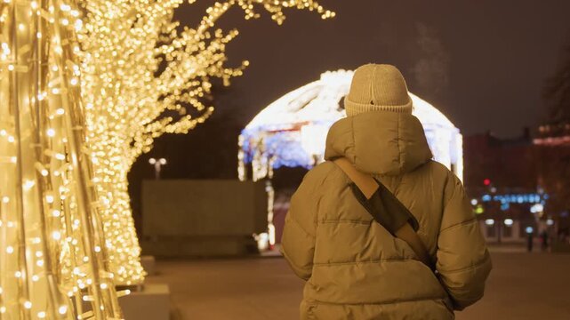 Lonely figure in illuminated park, solitary walker in heavy puffer coat pauses beneath golden tree lights, gazing at glowing white gazebo across plaza, soft bokeh highlights, cold breath visible,