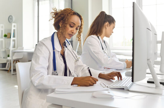 Doctors team at computer in clinic. Focused physicians with stethoscopes work at monitors, writing notes and updating records in a bright modern office. Clear concept of medical collaboration.