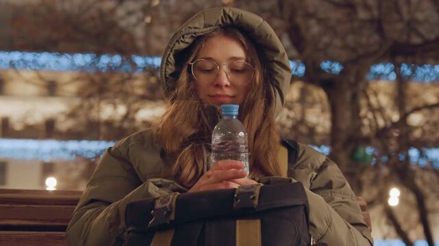 Girl resting outdoors. Caucasian girl enjoying winter moment. Young woman in park during peaceful snowy evening. Sitting on bench with water bottle amidst snowy winter evening lights
