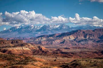 Fototapeta premium Red rock formations under snow-capped mountains, vast desert landscape, cloudy sky