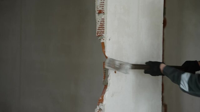 Close-up view of construction worker wearing protective mask and gloves using heavy sledgehammer to break through brick wall plaster during home renovation project