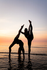 Fototapeta premium Two young women stretch in the water at sunset. A silhouette of dancers in gymnastic poses on the seashore. A concept of nature, sport, and pleasure.