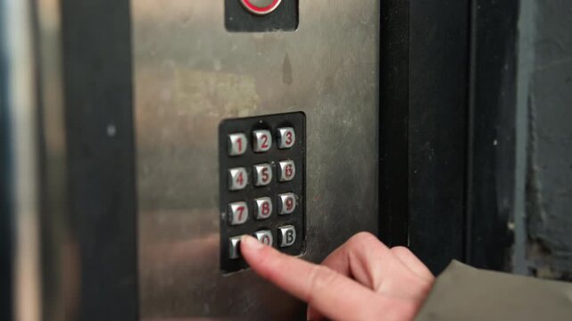 Finger entering building access code, resident presses metal keypad in dim lobby, closeup on numeric panel and sleeve, buzzer and camera visible, tense nocturnal entry scene with cautious atmosphere