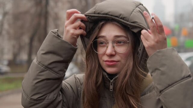 Young caucasian woman adjusting hood outdoors on cold city sidewalk, wearing glasses and puffer coat, subtle snow flurries dusting hair, hands moving to shield face, expressions shifting from pensive
