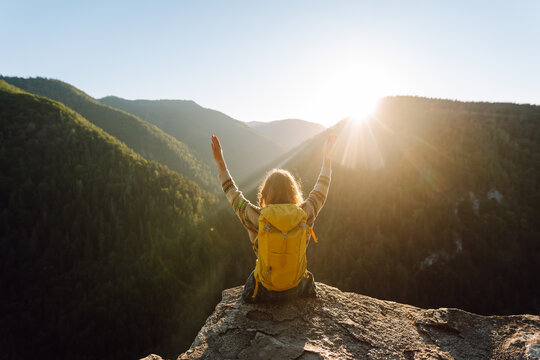 A female hiker with a backpack sits on the edge of a cliff at sunset. The young woman enjoys the mountain scenery outdoors. Concept of relaxation, hiking, and nature.