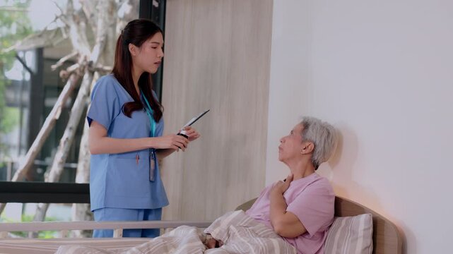 Young female nurse in blue uniform holding clipboard having caring conversation with anxious elderly woman patient resting on hospital bed showing medical report providing empathetic support