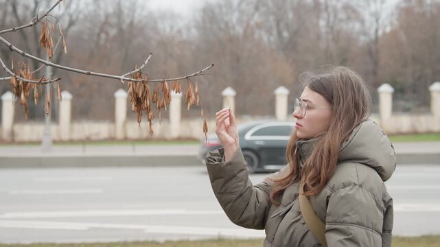 Medium shot woman in puffer jacket inspecting tree pods by busy roadside, glasses and backpack visible against moving traffic, candid city atmosphere with muted winter palette and thoughtful pause