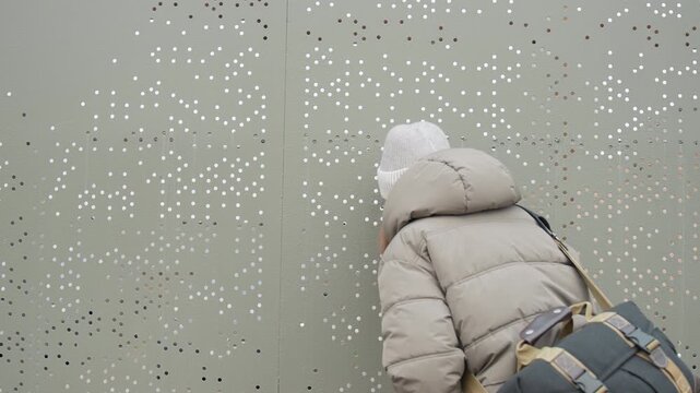 Caucasian woman explores perforated metal facade, walking along city wall, knitted hat and heavy coat, hand touching patterned holes, satchel on shoulder, brisk winter mood, architectural