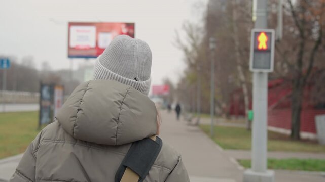 White woman walking on sidewalk near crosswalk, commuter paused at red pedestrian signal in overcast urban morning puffer jacket, knitted beanie and shoulder bag convey quiet routine and reflective