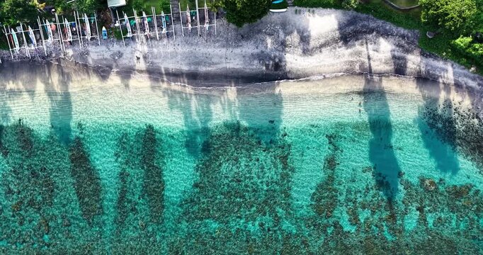 Top-down aerial drone footage of vibrant turquoise coral reef waters meeting the volcanic black sand shoreline in Amed, Bali, Indonesia with traditional fishing boats and swimmers along the tropical b