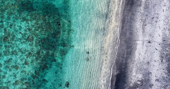Overhead aerial drone shot of swimmers enjoying crystal-clear turquoise water beside the volcanic black sand beach and coral reef coastline in Amed, Bali, Indonesia.
