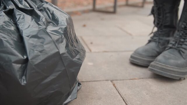 Black trash bag on sidewalk closeup showing heavy boots nearby, plastic sack crumpled on paved pavement, gritty urban atmosphere with scattered litter, low angle reveal of approaching footsteps