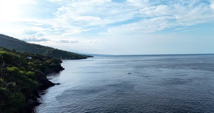 Drone aerial panorama of the peaceful coastline in Amed, Bali, Indonesia featuring calm ocean waters, traditional Balinese fishing boats, lush green hills, and dramatic tropical skies.