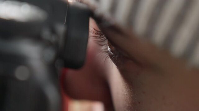 Tight eye portrait through camera, focused photographer peering through eyecup, soft skin tones, beanie shadow, concentrated composition, cinematic grain, natural lighting, creative direction, shallow