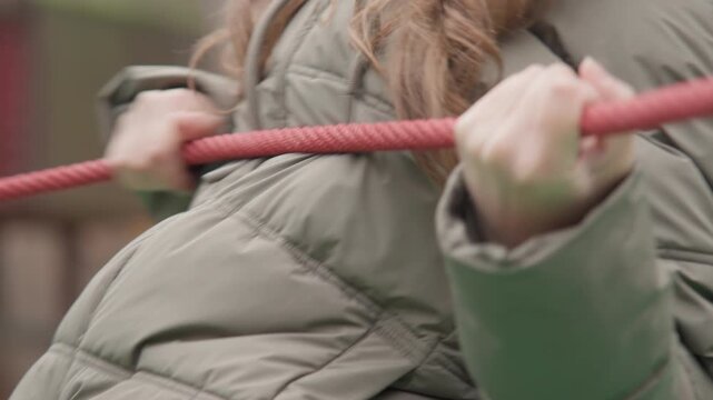 White woman gripping red rope playground, winter jacket beanie closeup hands pulling with tense knuckles, park background with muted autumn tones, candid training vibe showing resilience and teamwork,