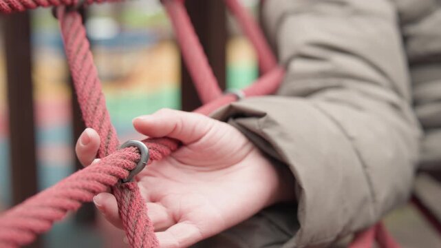 Child hand touching red rope net, winter coat sleeve, soft bokeh background, fingers exploring knot and weave, tactile curiosity captured in closeup, muted color palette, outdoor playground