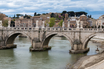 Fototapeta premium View on stone-arched bridge on Tiber river in Rome, balustrade, marble statues, cloudy sky, ochre and terracotta buildings, domes and umbrella pines rise on gently sloping Vatican hill
