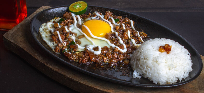 close up photo of spicy pork mask sisig with fried rice meal on a sizzling plate with egg and lemon drizzled with mayonnaise.