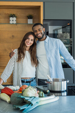 Diverse couple smiling at camera while embracing and preparing fresh vegetables for a healthy meal in their modern kitchen, symbolizing teamwork, happiness, and a healthy lifestyle at home