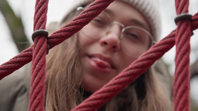 Playful white girl making faces behind rope net, tongue out and wry smile, glasses and beanie catching light, vlogger friend teasing camera with spontaneous reactions, candid winter park energy