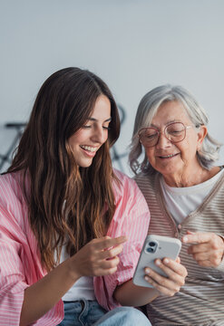 Young woman helping an older woman understand and use a smartphone, bridging a generational gap through technology and fostering family connection while smiling indoors