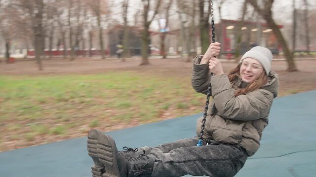 Woman riding zipline across playground, full swing motion captured, legs extended and laughing face, wooden platform and rope chain visible, overcast autumn park, playful adult enjoying ride