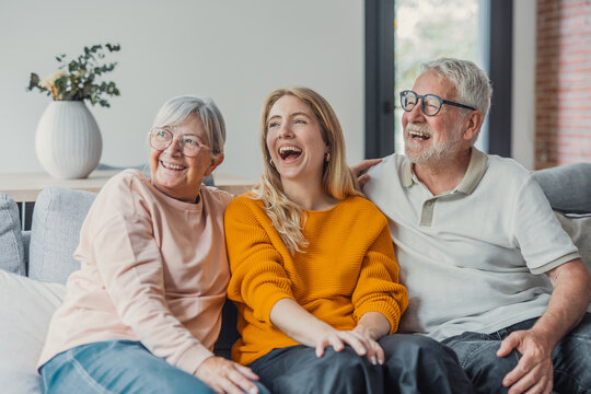 Three generations of a happy family, including a young woman and her senior parents, sitting together on a comfortable couch in their living room, sharing a joyful moment and laughing