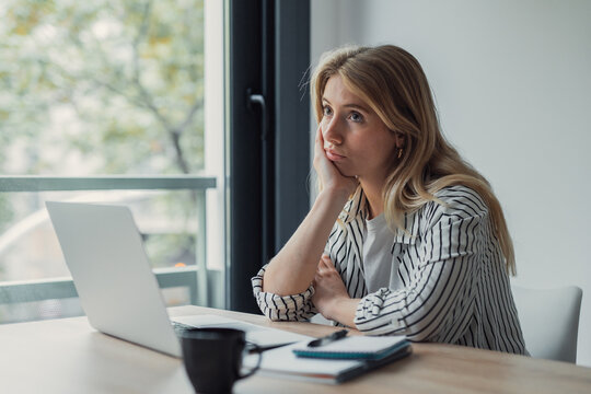Young woman in striped shirt procrastinating while working from home, gazing out the window from a laptop-strewn desk, thoughtful and unmotivated during remote study or workday
