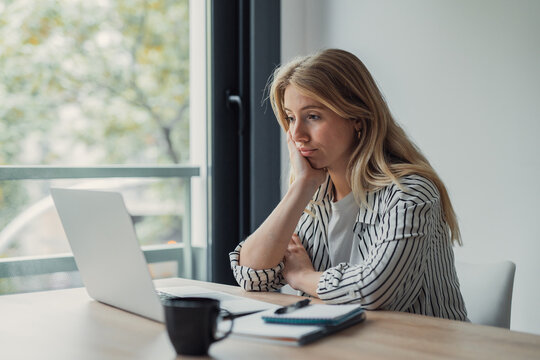 Young woman leaning on her hand, looking disengaged in front of her laptop during remote work, experiencing boredom, fatigue, and lack of motivation, possibly procrastinating