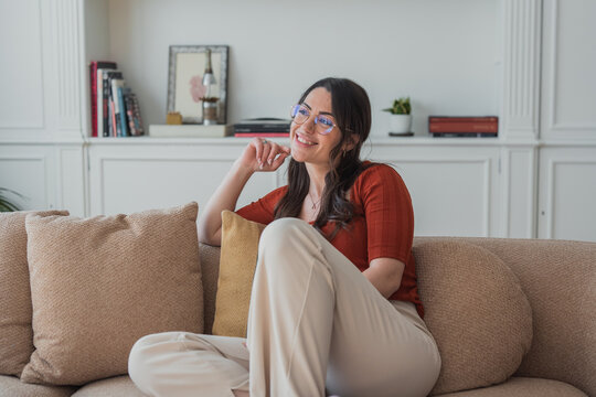 Smiling woman wearing glasses sitting comfortably on a sofa in her modern living room, enjoying a moment of peaceful contemplation and a relaxed lifestyle at home