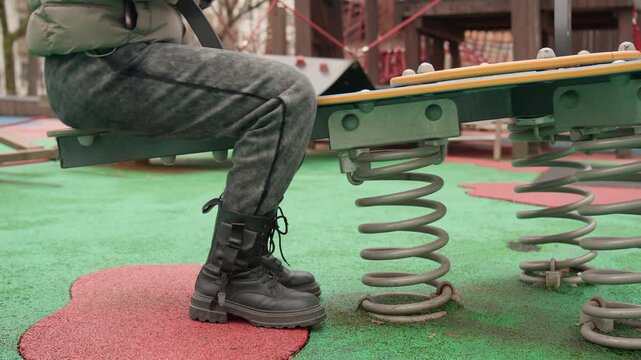Closeup boots on playground spring seat showing adult testing bounce on green rubber surface with metal coil suspension, gray pants, black lace boots pressing down, urban park atmosphere, textured