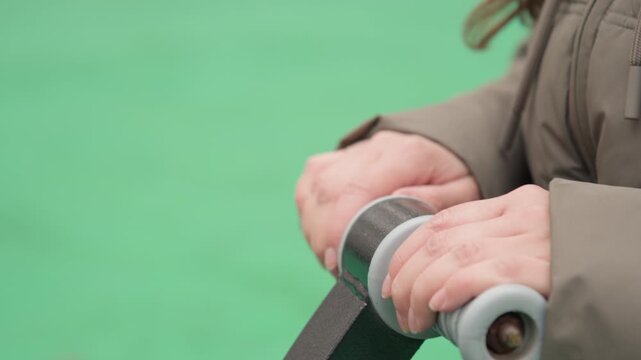 Closeup hands gripping playground handle in cold weather, adult in puffer jacket holding textured roller handle, subtle wrist movement and steady pressure on metal bar, calm urban park scene