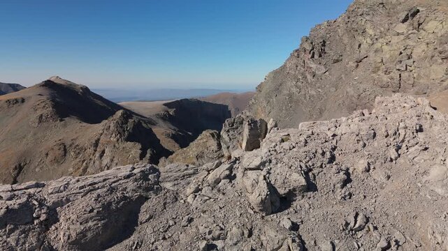 Drone footage captures mountain goats bounding along a barren ridge through harsh alpine terrain, with dramatic rocky cliffs, steep slopes and clear blue sky above distant mountain peaks