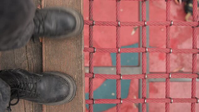 Closeup boots over red safety net wooden plank gap reveals turquoise water below, worn leather soles and scuffed laces, steady stance and cautious step, mesh pattern frames vertical drop, tense POV