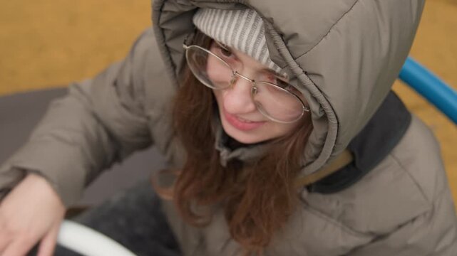 Young woman in puffer jacket playing on playground carousel, smiling while gripping white wheel wearing hooded coat, knit beanie and glasses chilly overcast afternoon, candid lifestyle broll capturing
