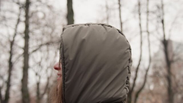 White woman profile in park smiling and surprised, hooded parka and beanie playful hand gesture and laughter, bare trees and crisp air, candid reaction shot conveying joyful discovery and youthful