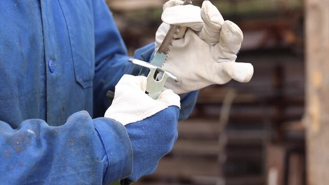 A worker in protective gloves replaces the blade on a sabre saw. A craftsman in a blue work uniform and strong protective gloves installs a saw blade into the chuck of an electric sabre saw
