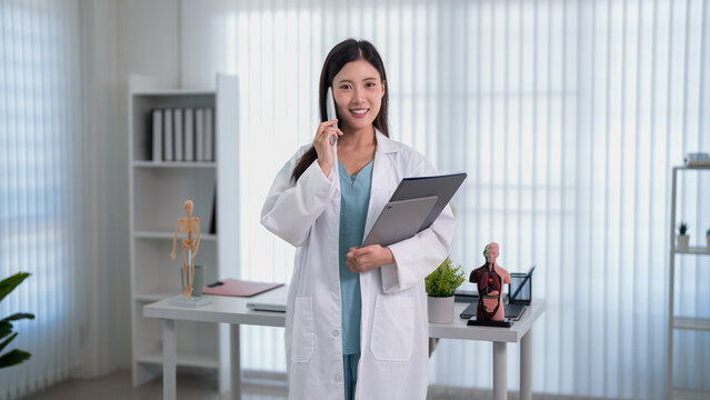 young Asian female doctor talking phone holding medical chart patient consultation telehealth healthcare professional modern clinic office bright natural light