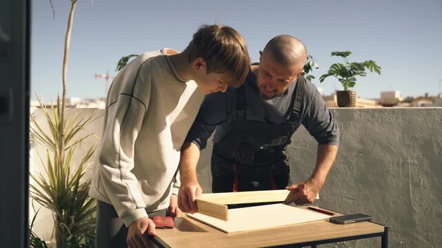 Craftsman father teaching his teenage son woodworking skills on sunny rooftop terrace, demonstrating how to build a wooden frame and sharing practical craftsmanship knowledge and family traditions