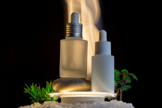Mystical night spa cosmetics mockup. Two blank frosted glass dropper bottles on zen stones and sea salt. Glowing warm orange light and smoke effect from a humidifier on a black background with copy sp