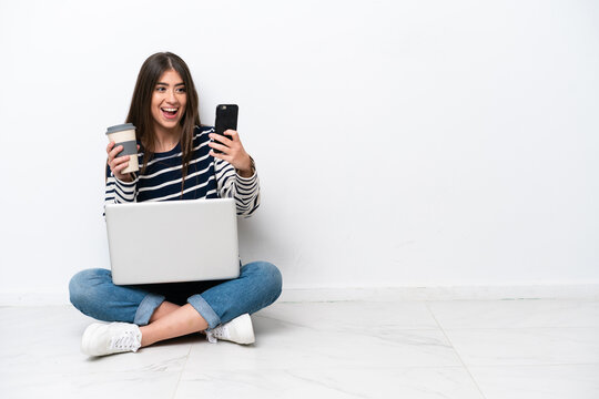 Young caucasian woman with a laptop sitting on the floor isolated on white background holding coffee to take away and a mobile