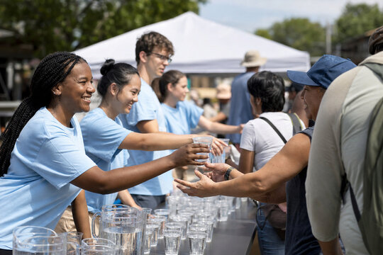 Several people stand at a table full of cups filled with water. They hand out water to others in an outdoor space. The sun shines and trees provide some shade.